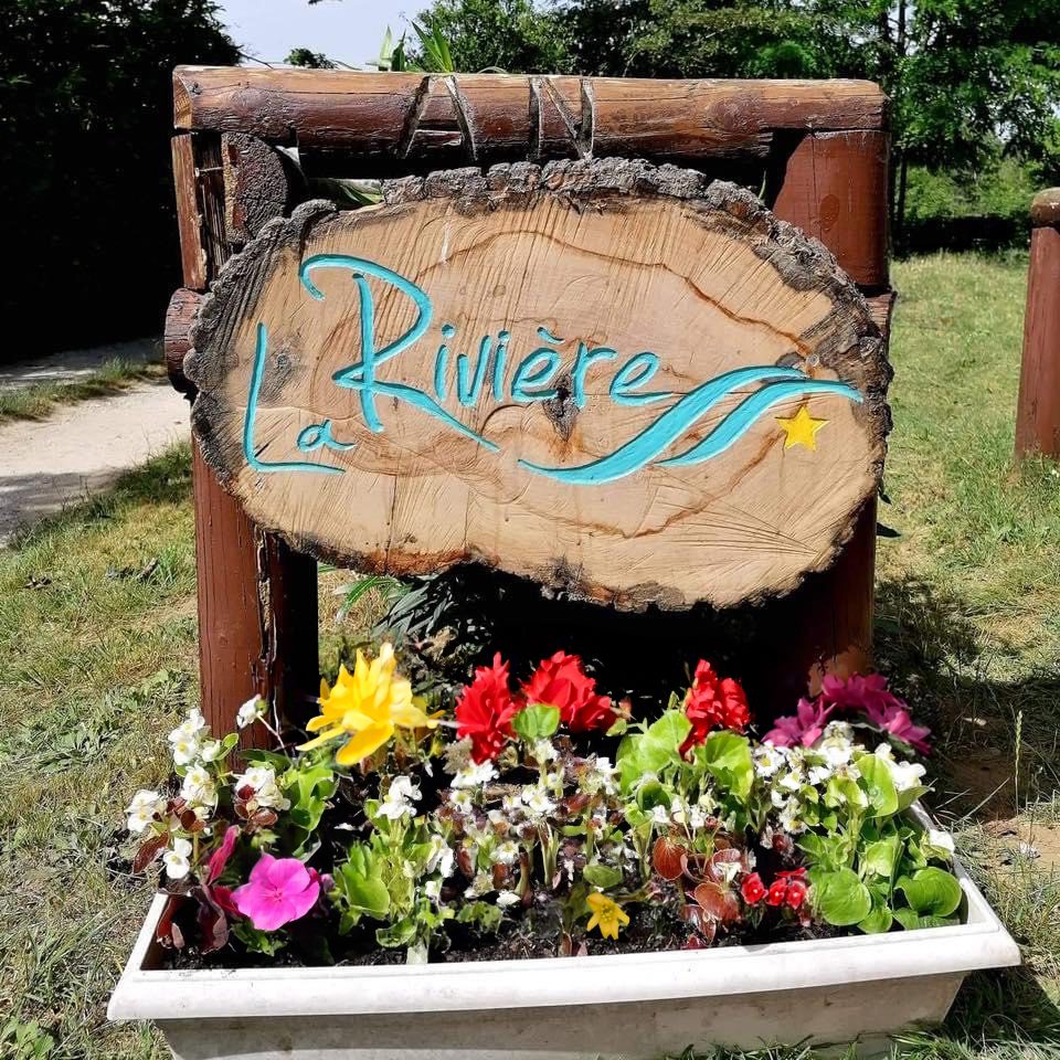 Sculpture en bois avec inscription "La Rivière" et fleurs colorées en avant-plan.